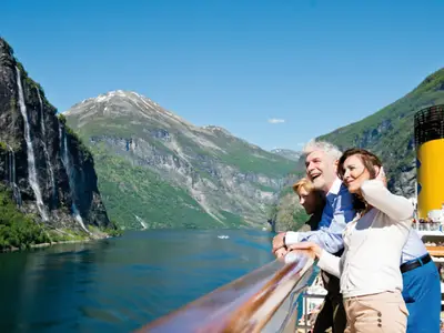 Drei Passagiere der Costa Fortuna auf Deck blicken auf den norwegischen Geirangerfjord. Eindrucksvolle Wasserfälle von grünen Bergen, blauer Himmel, glückliche Gesichter am Deck.