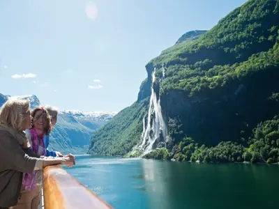 Mehrere Passagiere der Costa Fortuna stehen am Deck und blicken auf einen atemberaubenden Fjord mit hohen Bergen und Wasserfällen.