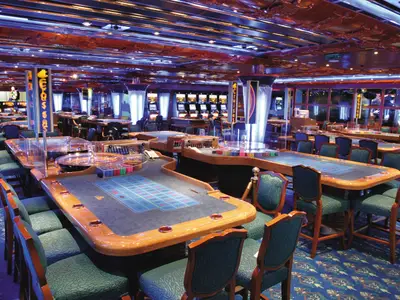 Innenansicht des Casino Neptunia an Bord der Costa Fortuna mit Roulette- und Kartenspieltischen. Die Stühle sind leer, die Decke kunstvoll. Im Hintergrund Slotmaschinen.