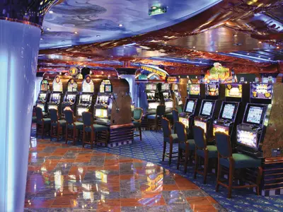 Das Casino Neptunia an Bord der Costa Fortuna. Reihen beleuchteter Slotmaschinen mit Bildschirmen und Stühlen. Der Boden ist teils Teppich, teils glänzende Fliesen. Die Decke ist kunstvoll dekoriert, blaue Lichter schaffen eine lebhafte Spielatmosphäre.