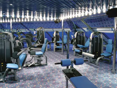 Ein geräumiges Fitnesscenter an Bord der Costa Fortuna mit modernen Trainingsgeräten in Grau und Blau. Der Raum hat eine blaue Decke mit hellen Lichtern, große Fenster und einen gemusterten Teppich. Keine Personen sichtbar.