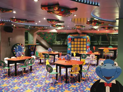 Innenansicht des Squok Clubs an Bord der Costa Fortuna mit bunten Kindertischen und Stühlen. Der Raum ist dekoriert mit Leuchten, Spielelementen und einem Memory-Spielbrett. Ein großer blauer Drache schmückt den Raum.