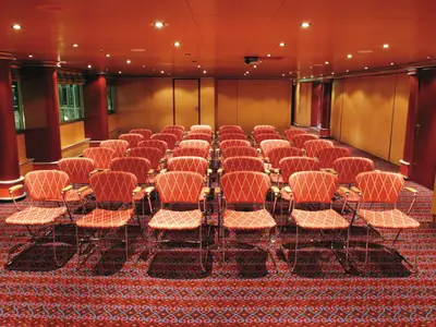 Blick in den Konferenzraum Saturnia an Bord der Costa Fortuna. Der Raum ist mit roten Teppichboden und zahlreichen orangefarbenen Stühlen in Reihen ausgestattet.