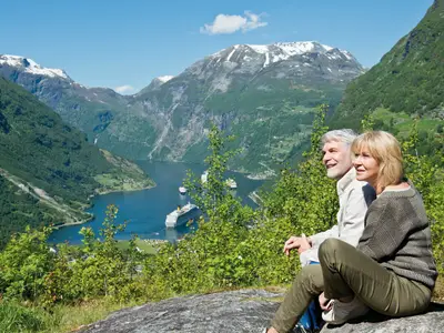 Ein lächelndes Paar blickt auf den norwegischen Geirangerfjord. Unten fährt die Costa Fortuna, umgeben von grünen Bergen und schneebedeckten Gipfeln.