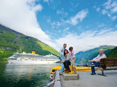 Die Costa Fortuna im norwegischen Geirangerfjord. Eine Gruppe Reisender blickt vom Kai auf das Schiff, umgeben von gruenen Bergen unter blauem Himmel.