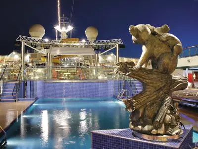 Der beleuchtete Poolbereich an Bord der Costa Fortuna bei Nacht. Eine bronzene Skulptur zeigt einen Koala mit Jungem auf einem Ast. Im Hintergrund sind die hell erleuchteten Decks und Schiffsaufbauten zu sehen.