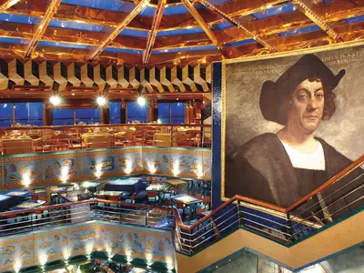 Innenansicht des mehrstufigen Restaurants Cristoforo Colombo an Bord der Costa Fortuna. Oben Tische mit gelben Tischdecken, unten blaue Sitzgruppen und ein riesiges Christoph Kolumbus Gemälde.