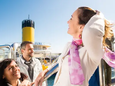 Drei lächelnde Passagiere genießen die Sonne auf dem offenen Sonnendeck der Costa Fortuna. Im Hintergrund ist der gelbe Schornstein des Schiffes und das weite Meer zu sehen.