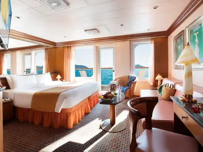 Helle Suite mit Balkon und Meerblick an Bord der Costa Fortuna. Sie zeigt ein Doppelbett, Sofa, Tisch mit Früchten und Champagner sowie einen Arbeitsbereich.