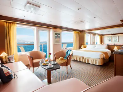 Eine helle Suite an Bord der Costa Fortuna mit grossen Fenstern zum Meer. Das Zimmer hat ein Doppelbett, einen kleinen Esstisch mit zwei Stuellen und eine Couch. Mehrere Bilder schmuecken die Waende.