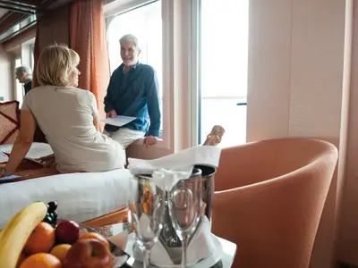 Ein Paar entspannt in einer Suite an Bord der Costa Fortuna. Die Frau sitzt auf dem Bett, der Mann lächelt am Balkonfenster. Champagner und Obst stehen bereit.