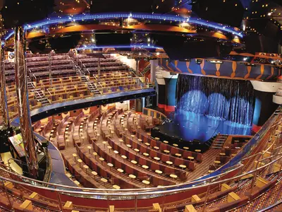Weitwinkelaufnahme des Theaters Rex an Bord der Costa Fortuna. Die Bühne mit blauem Vorhang und die leeren Zuschauerränge mit Sitzreihen und Tischen sind gut zu sehen. Blaue Lichter schmücken die Decke.