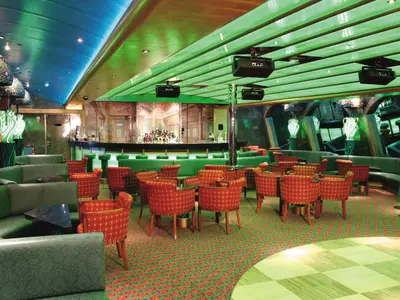 Ansicht der stilvollen Bar-Lounge Conte Verde an Bord der Costa Fortuna. Sie zeigt grüne Polster, rote Sessel und eine beleuchtete Bar mit Flaschen.