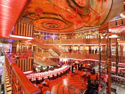 Das lebhafte Atrium Welcome der Costa Pacifica. Eine beleuchtete zentrale Bar, elegante geschwungene Treppen und orangefarbene Sitzbereiche füllen den mehrstöckigen Raum. Rote Musiknoten dekorieren die goldene Decke. Zahlreiche Gäste und Crew sind im warm beleuchteten Innenbereich zu sehen.