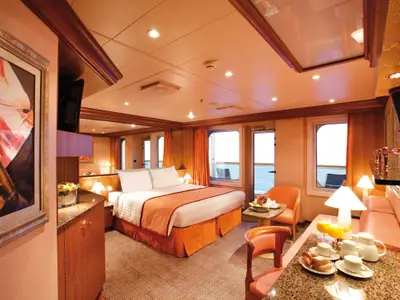 Großzügige Suite an Bord der Costa Pacifica mit Doppelbett, Sitzbereich, Balkontür zum Meer und serviertem Frühstück im Vordergrund.