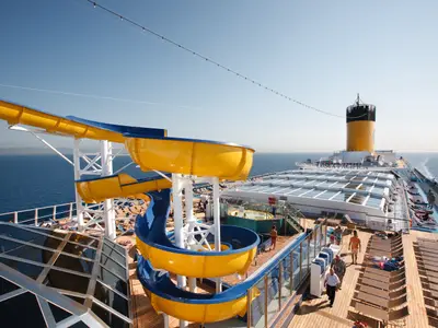 Blick auf die spiralförmige gelbe Wasserrutsche auf dem Sonnendeck der Costa Pacifica, dahinter Liegestühle, Passagiere und der gelbe Schornstein.