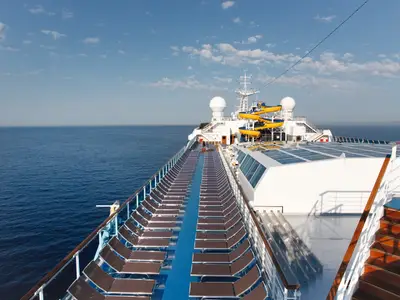 Blick über das weitläufige, sonnige Sonnendeck an Bord der Costa Pacifica mit blauen Liegen, einer Wasserrutsche und Blick auf das blaue Meer.