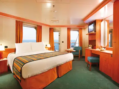 Blick in eine helle Balkonkabine der Costa Pacifica. Ein großes Bett, Nachttisch und ein Schreibtisch mit TV sind zu sehen. Die Balkontür und Fenster bieten eine weite Aussicht auf das Meer.
