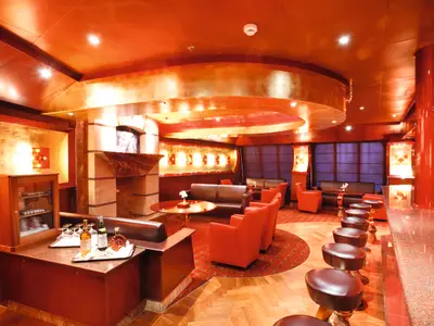 Der Innenbereich der Classico Amapola Bar an Bord der Costa Pacifica mit roten Sesseln, dunklen Sofas, einer Bar und einer Auswahl an Spirituosen. Die Decke ist goldfarben, und überall sorgen Lichtinstallationen für eine gemütliche Atmosphäre.