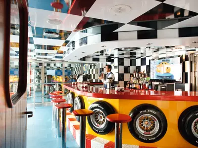 Barkeeper mixt in der Racing-Bar Scuderia Costa der Costa Pacifica. Die Bar mit Rennsport-Thema: Schachbrettmuster, Reifen, rote Tresen. Ein TV zeigt Rennen.