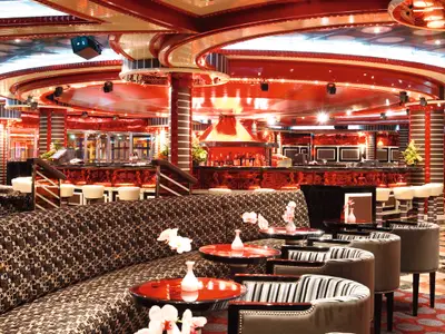 Die stilvolle Around the clock Lounge an Bord der Costa Pacifica. Eine luxuriöse Bar mit roter Decke, Säulen in rot-weiß-schwarz und gemusterten Sofas. Die Bar ist mit Flaschen bestückt, und es gibt mehrere Tische mit Blumenvasen.