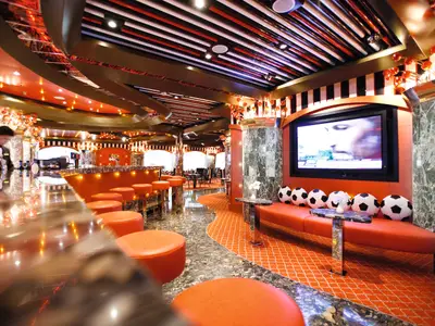 Moderne Sportbar "Route 66" an Bord der Costa Pacifica. Orangefarbene Barhocker, Theke, TV-Bildschirm mit Fußballkissen und ein Barkeeper.