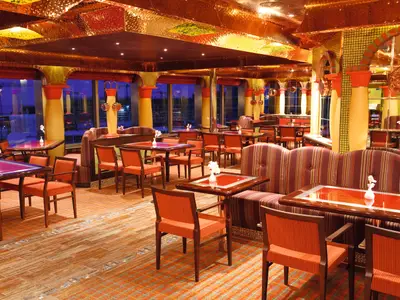 Das La Paloma Restaurant der Costa Pacifica. Viele orange Stühle und Bänke an dunklen Holztischen füllen den opulenten Raum. Goldene Decke und große Fenster zur abendlichen Skyline.