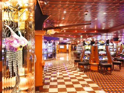 Geräumiges Casinò Flamingo an Bord der Costa Pacifica mit Reihen von leuchtenden Spielautomaten, Hockern, gemustertem Boden und einer auffälligen, bunten Kristall-Lichtinstallation.