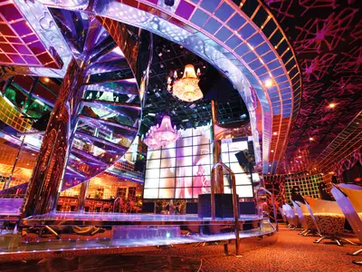 Ein weitläufiger Blick auf die futuristische Disco Fever an Bord der Costa Pacifica. Glänzende, chromfarbene Oberflächen, bunte LED-Lichter und große Kronleuchter prägen den Raum. Eine Bühne und Sitzgelegenheiten sind zu sehen, alles ohne Personen.