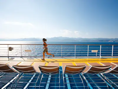 Eine Frau joggt auf dem sonnigen Oberdeck der Costa Pacifica. Im Hintergrund das Meer und Berge. Das Deck ist blau-gelb mit Sonnenliegen vorn. Ein klarer Tag.