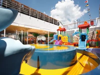 An Bord der Costa Pacifica: Heller Kinderbereich mit farbenfrohen Wasserspielplatz-Elementen, Rutschen und Klettergerüsten auf dem offenen Deck. Im Hintergrund ist eine Küstenstadt zu sehen. Blauer Himmel mit Wolken.