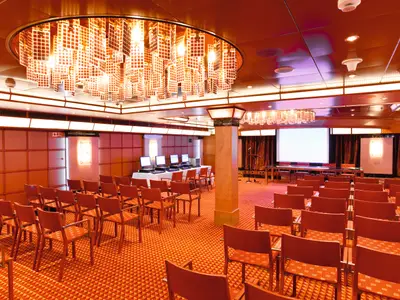 An Bord der Costa Pacifica: Der Amadeus Konferenzraum mit oranger Bestuhlung, Computertischen und Projektionsfläche. Eine moderne Deckenleuchte dominiert den Raum.