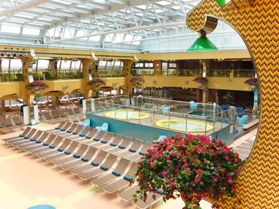 Der helle Lydo Calypso Poolbereich an Bord der Costa Pacifica mit Liegestühlen, zwei Jacuzzis und umlaufenden Restaurantebenen unter einem Glasdach. Eine bunte Mosaikwand ist rechts sichtbar.
