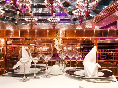 Nahaufnahme eines elegant gedeckten Tisches im Club Blue Moon Restaurant an Bord der Costa Pacifica. Gläser, Teller, Besteck und gefaltete Servietten sind zu sehen. Im Hintergrund schimmernde kupferfarbene Strukturen und Deckenleuchten. Eine einzelne weiße Orchidee ziert den Tisch.