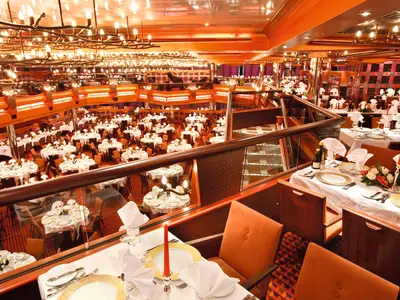 Grosszügiger Blick auf das mehrstöckige Hauptrestaurant New York New York an Bord der Costa Pacifica. Festlich gedeckte Tische mit weißer Tischwäsche, roten Kerzen und Blumen. Warme Beleuchtung.