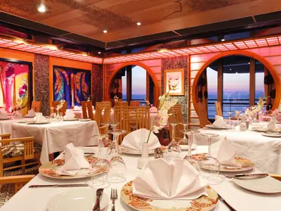 Gedeckte Tische im asiatischen Samsara Restaurant an Bord der Costa Pacifica mit Rundfenstern zum Meer bei Sonnenuntergang.