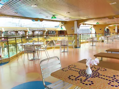 Blick in ein buntes Restaurant an Bord der Costa Pacifica. Zahlreiche Tische mit silbernen Stühlen stehen auf einem farbigen Boden. Gelb gemusterte Wände und große Fenster zum Deck prägen den Raum. Weiße Orchideen schmücken einen Tisch im Vordergrund.