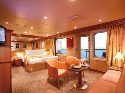 Eine elegante Grand Suite an Bord der Costa Pacifica. Grosses Bett, zwei Sessel, Sofa, Tisch mit Obst und Champagner, sowie Balkon mit Meerblick.