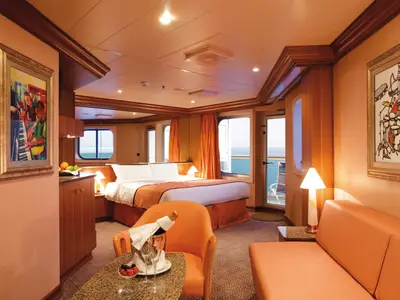 Eine helle, geräumige Suite an Bord der Costa Pacifica mit grossem Bett, orangefarbenem Sofa und Sessel. Durch die bodentiefen Fenster ist das Meer sichtbar. Dekoriert mit Kunstwerken.