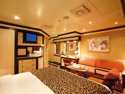 Geräumige Suite an Bord der Costa Pacifica mit hellem Interieur, großem Bett, beigem Schrank, rotem Sofa und Essbereich mit Tisch und Stühlen.