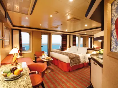 Geräumige Suite an Bord der Costa Pacifica mit Doppelbett, orangefarbenen Möbeln, einem Schreibtisch mit Obstschale und Fenstern sowie einer Balkontür mit Meerblick.