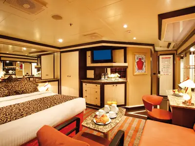 Ansicht einer geräumigen Suite an Bord der Costa Pacifica mit großem Doppelbett, Spiegelwand, TV-Bereich und einem gemütlichen Frühstückstisch mit Gebäck und Säften.