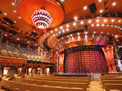 Das Stardust Theater an Bord der Costa Pacifica zeigt eine aufwendige Bühne mit rotem Vorhang und leere Sitzreihen. Ein bunter Kronleuchter und Notenschlüssel zieren den Raum.