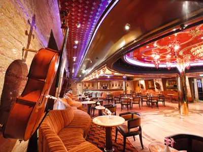 Eine luxuriöse Bar-Lounge an Bord der Costa Pacifica. Orangefarbene Sofas und Tische füllen den Raum, eine Bar mit Barkeeper ist im Hintergrund. Dekoriert mit Celli und Geigen, unter einer kunstvollen Decke.