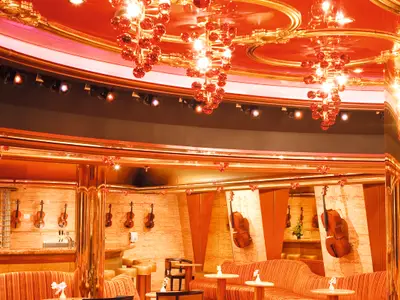 Opulenter Innenbereich an Bord der Costa Pacifica: Eine Bar-Lounge mit Wanddekoration aus Geigen und Cello, goldenen Säulen, glänzenden roten Kronleuchtern und Deckenakzenten.