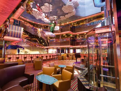 Das mehrstöckige Atrium an Bord der Costa Serena mit bunten Säulen, Treppen, Figuren an Wolken und einem gläsernen Aufzug.