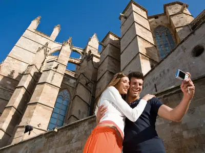 Ein junges Paar macht ein Selfie vor der historischen Kathedrale in Palma de Mallorca bei einem Landausflug der Costa Serena.