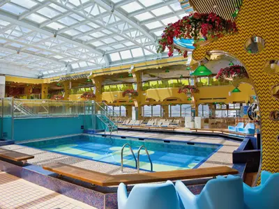Ein großer Swimmingpool unter einem Glasdach an Bord der Costa Serena mit gelb gemusterten Wänden und Holzliegen im Hintergrund.