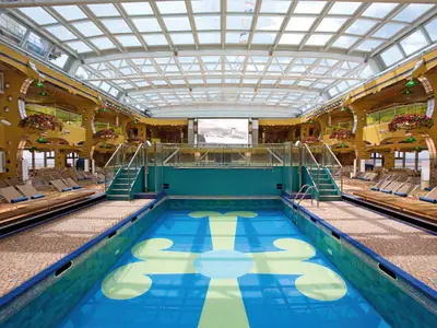 Ein blickgeschützter Poolbereich mit hellblauem Mosaikbecken, Sonnenliegen und einer großen Videoleinwand an Bord der Costa Serena.