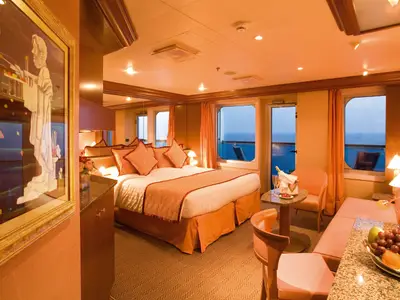 Einblick in eine großzügige Suite mit warmem Licht an Bord der Costa Serena, ausgestattet mit Doppelbett, Sitzecke, Obstkorb und Balkon zum Meer.